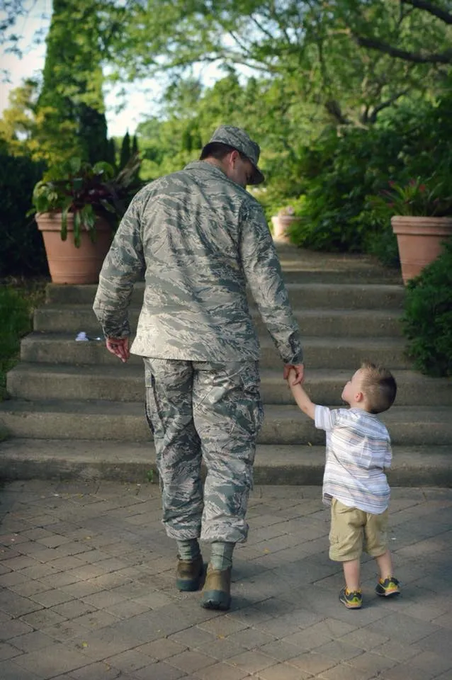 Service member in uniform holding his young son's hand on a brick path